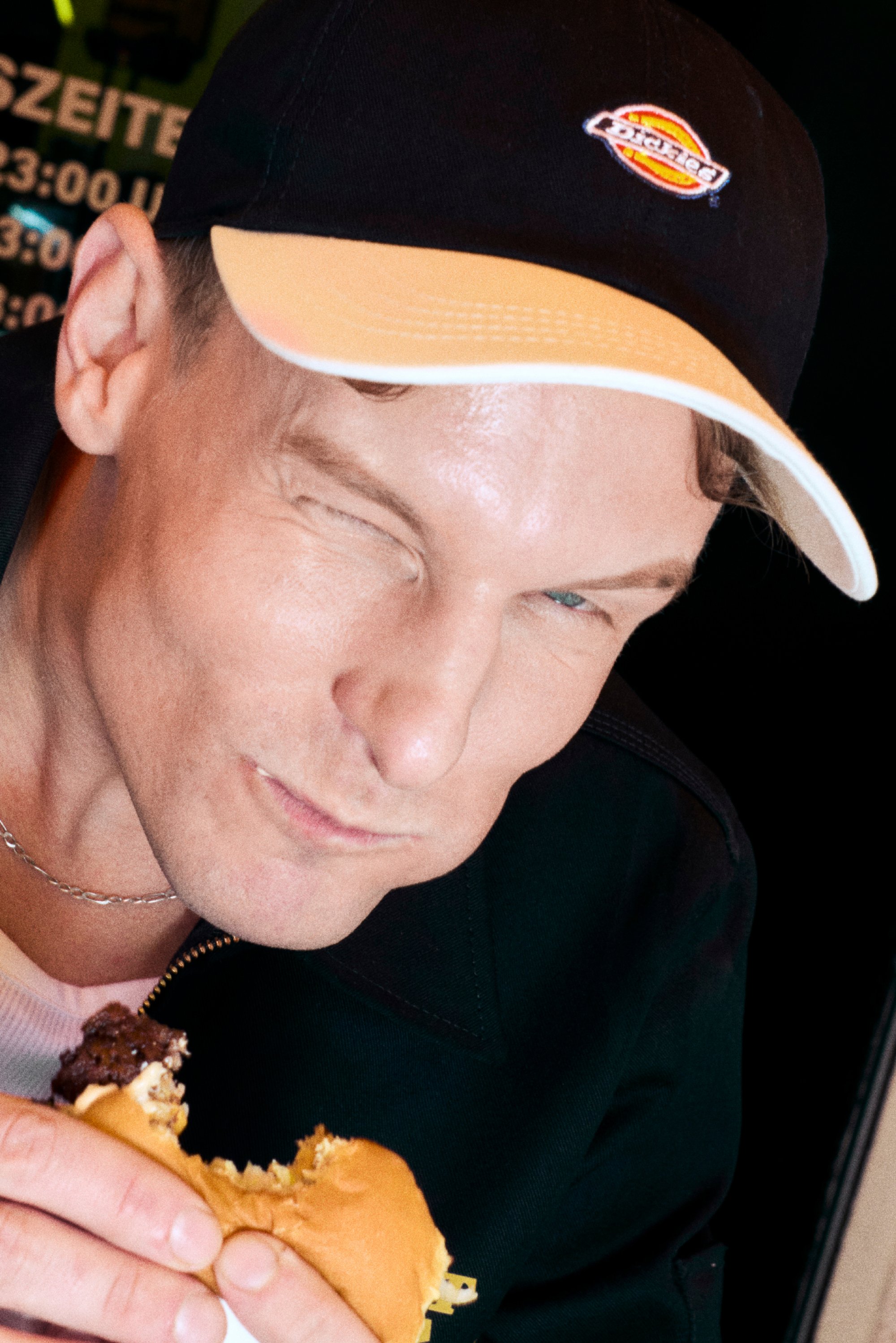 Ein Mann mit einer schwarz-orangefarbenen Kappe zwinkert, während er einen teilweise gegessenen Cheeseburger hält, mit einem konzentrierten, verspielten Ausdruck.