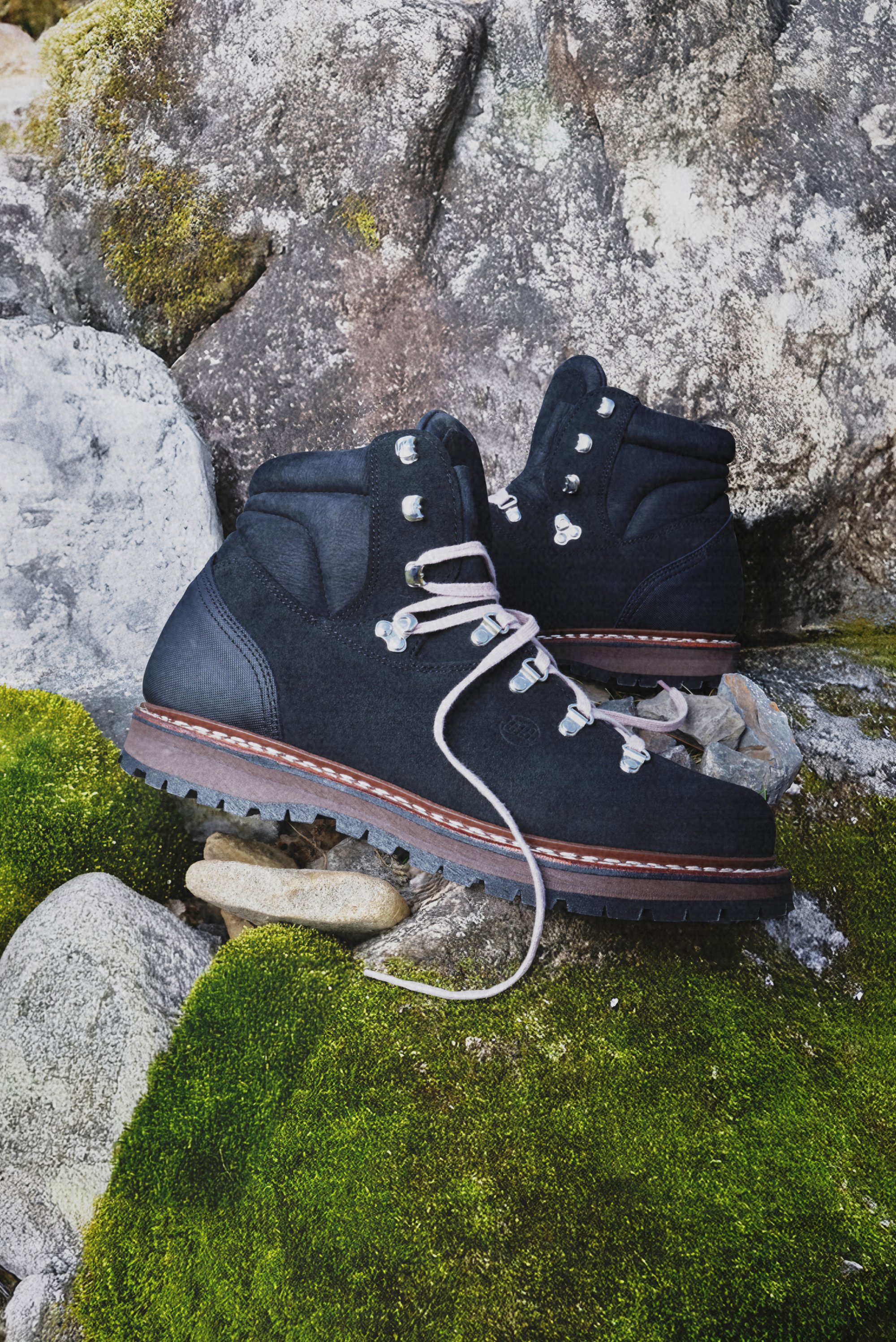Black hiking boots with silver eyelets and laces rest on green moss and grey rocks, showcasing a rugged outdoor setting.