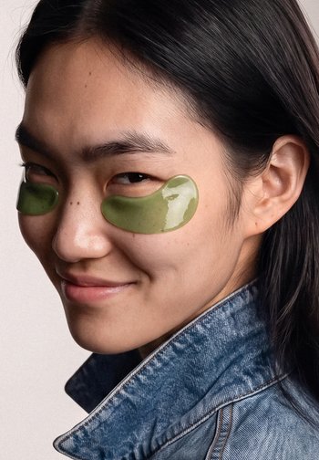 Jeune femme aux longs cheveux bruns portant des patchs sous les yeux verts en gel et une veste en jean, souriant légèrement vers la caméra.