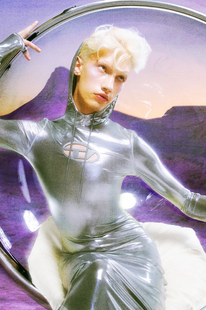 A figure with platinum blonde hair poses in a shiny, silver outfit inside a circular frame, against a purple and orange background.