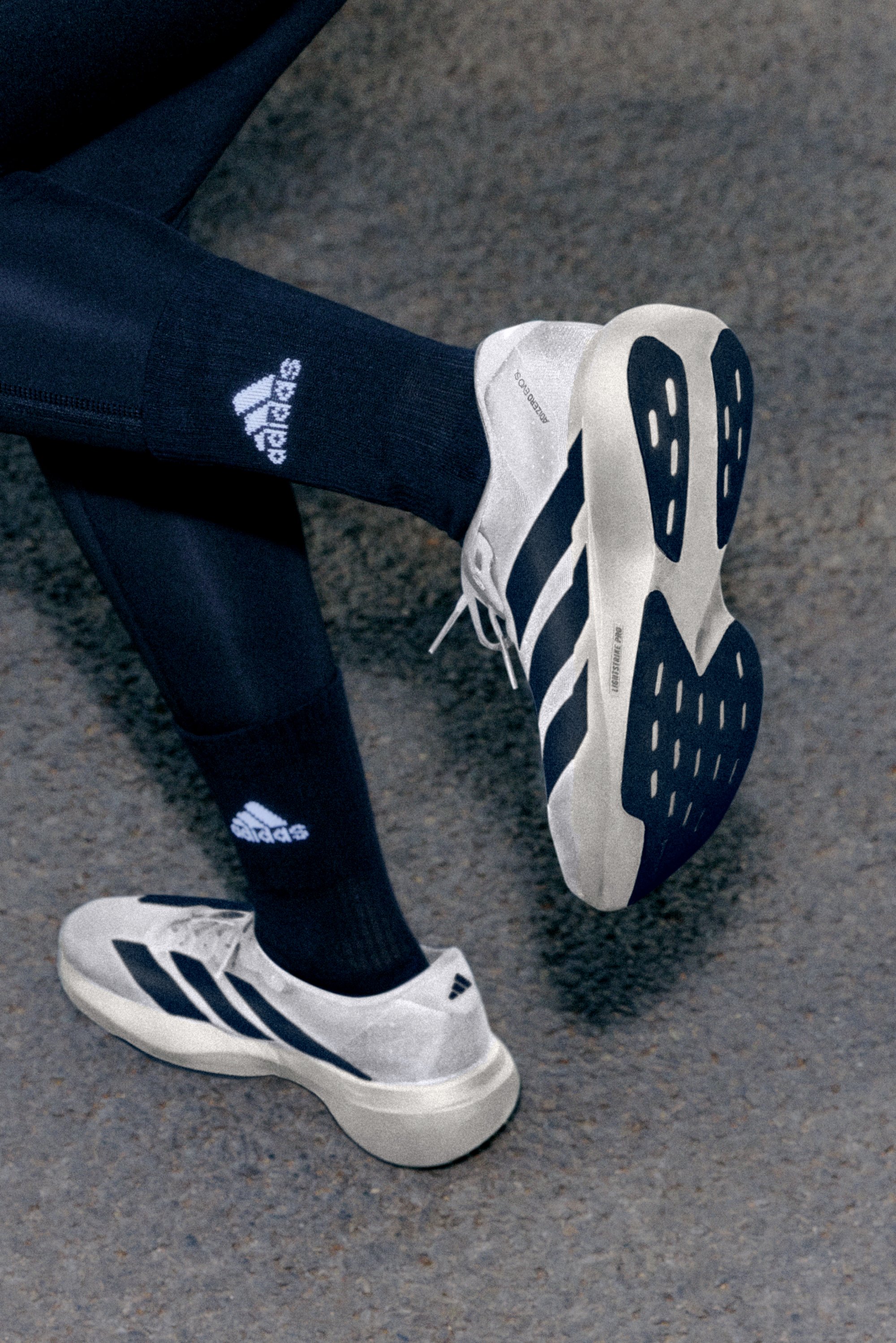 Primer plano de pies que llevan zapatillas Adidas en gris y azul marino, y calcetines negros con el logo de Adidas, sobre una superficie texturizada.