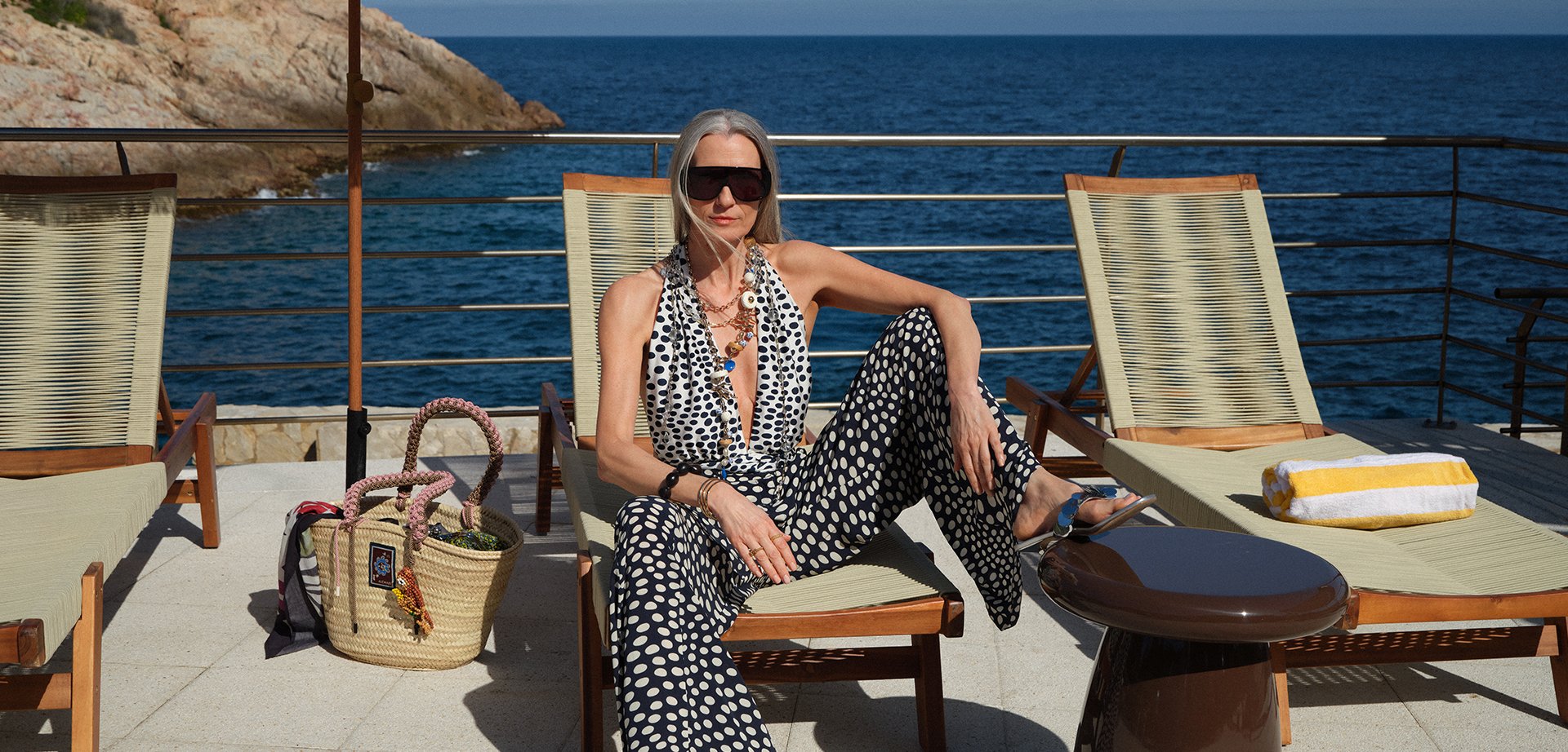 Frau in gemustertem Overall und Sonnenbrille sitzt entspannt auf einer Sonnenliege am Meer, zwei leere Liegestühle in der Nähe und eine Strohstrandtasche auf dem Boden.