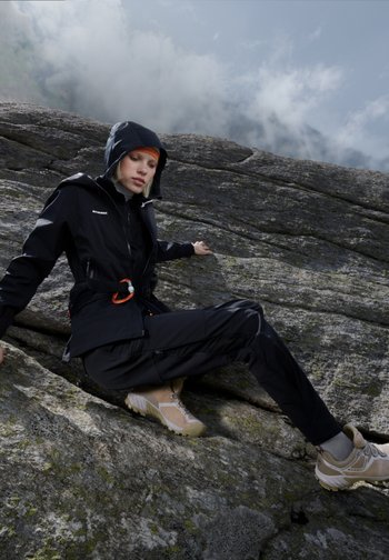 Ein Kletterer in schwarzer Outdoor-Ausrüstung sitzt auf einer felsigen Oberfläche und schaut nachdenklich in den bewölkten Himmel. Ein leuchtend oranges Stirnband bringt Farbe ins Bild.