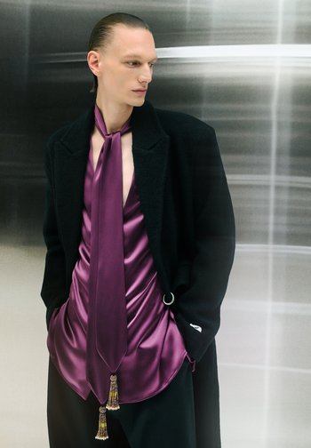 Black wool coat over a deep purple satin top with a tie. Top features draped texture and tassel details. Smooth, elegant silhouette.