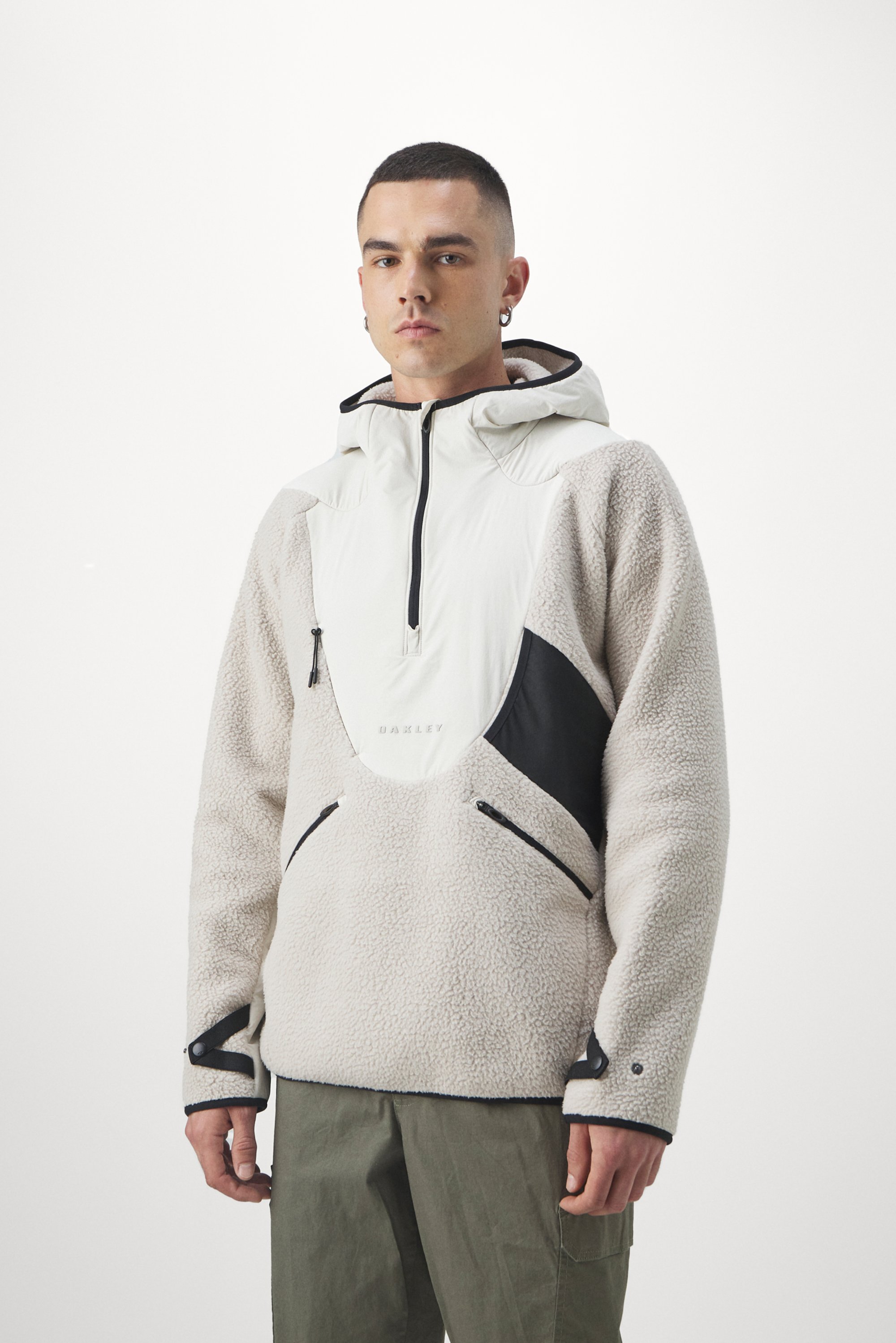 A young man stands facing forward, wearing a stylish beige and white fleece pullover with a hood, paired with olive green pants.