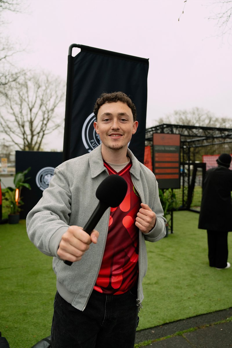 Young man in gray jacket and red shirt holds a microphone outdoors at an event with banners and greenery in the background.