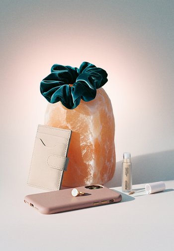 Turkusowa welurowa gumka do włosów na lampie solnej, beżowy etui na karty, różowy telefon ze złotym pierścieniem oraz otwarty korektor Kiko na białej powierzchni.