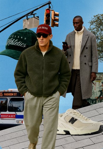 Two men are dressed in casual clothes. One wears a green fleece jacket and joggers, while the other dons a grey coat over a beige jumper. Notable accessories include a green cap and light trainers.