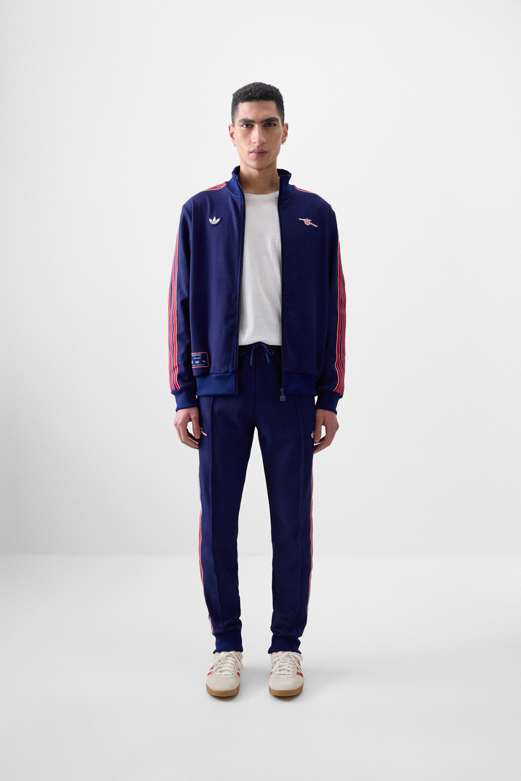 Young man stands in a minimalistic space, wearing a navy tracksuit with red stripes and white sneakers, hands relaxed at his sides.