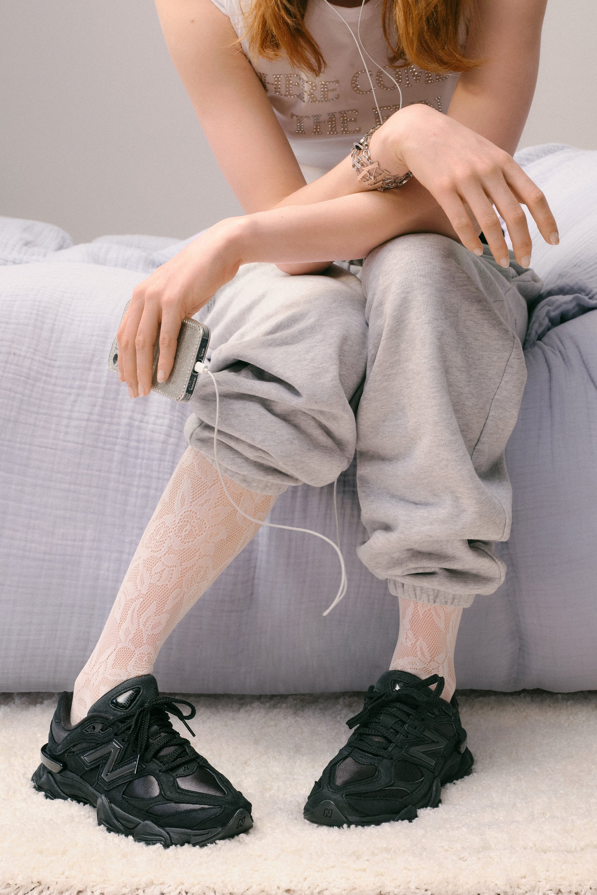 Una persona è seduta su un letto, indossando pantaloni grigi e scarpe da ginnastica nere, con un telefono scintillante e degli auricolari in mano, circondata da texture morbide.
