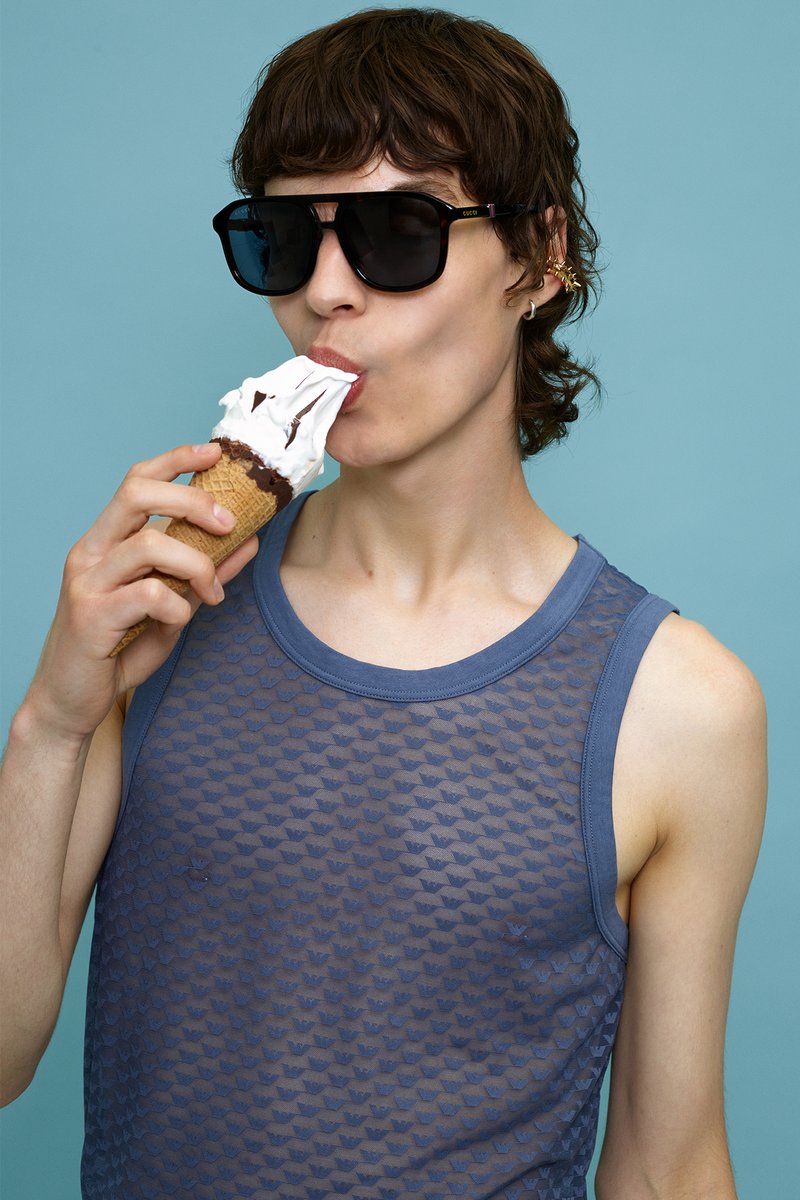 Iemand houdt een wafelcone vast met ijs, belegd met slagroom en chocoladesaus. Draagt een oversized zwarte zonnebril en een blauwe tanktop.