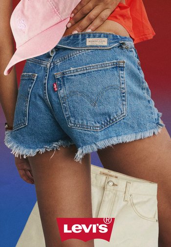 Persona che indossa pantaloncini Levi's in denim blu sfilacciati e un cappellino rosa, con una mano sulla vita, davanti a uno sfondo rosso e blu.