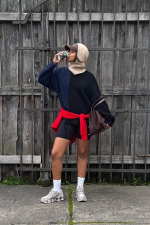 Übergroßer Strickpullover in Marineblau und Schwarz, mit rotem Taillengürtel, schwarzen Shorts, grauen Turnschuhen und einer beigen Strickmütze, die ein Getränk hält. Holzzaun im Hintergrund.
