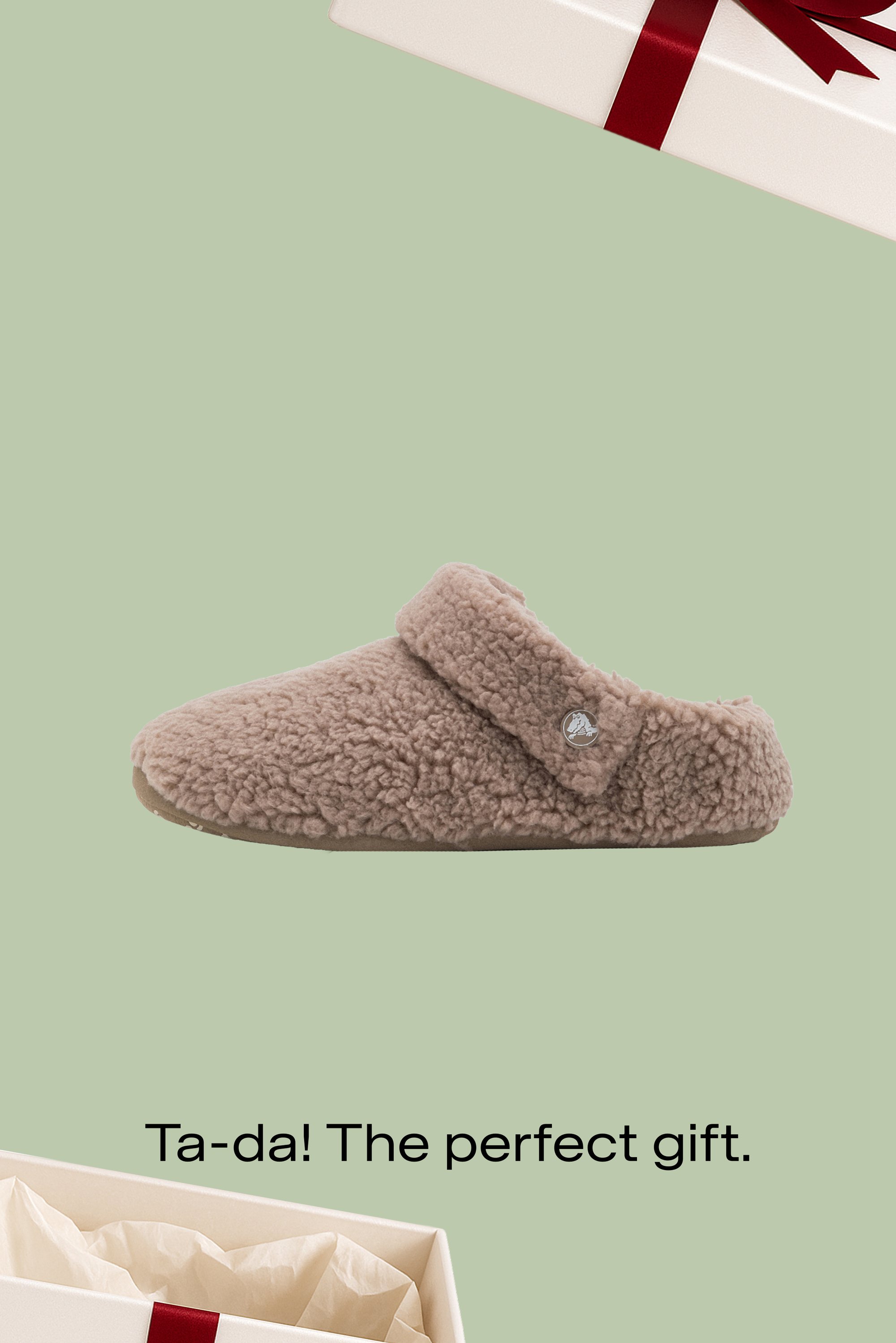 Brauner, pelziger Hausschuh zum Hineinschlüpfen mit verstellbarem Riemen vor grünem Hintergrund, daneben eine teilweise geöffnete Geschenkbox mit rotem Band.