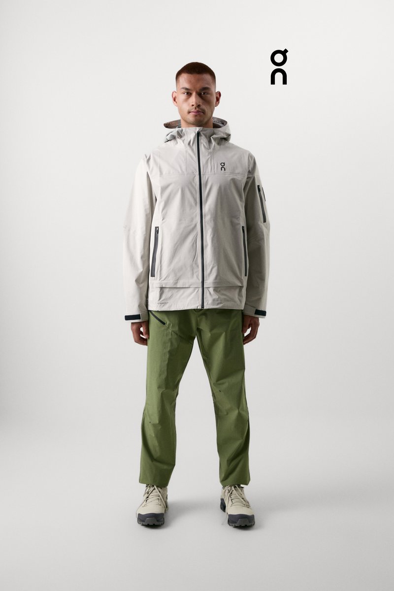 A young man stands confidently, wearing a light grey jacket and green trousers, against a minimalistic background, showcasing a stylish athletic look.