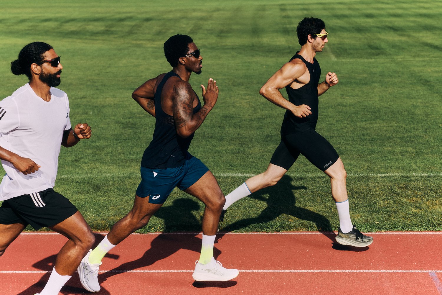 Three male runners in athletic gear sprint on a track, with green grass in the background. Sunlight enhances their focused expressions.