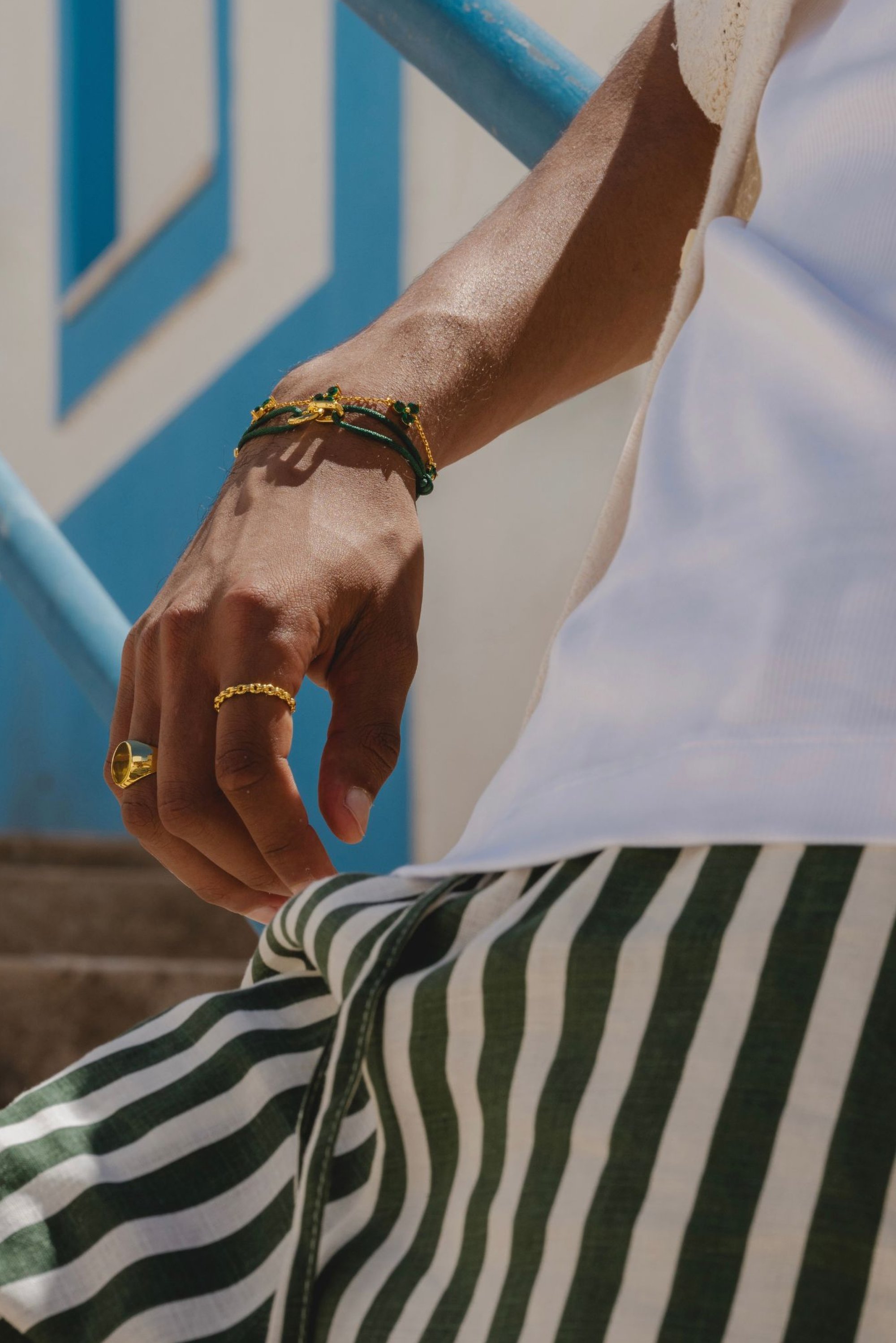 Anillo de oro y pulsera con acentos verdes en la mano; llevando una camisa blanca y pantalones a rayas verdes y blancas.