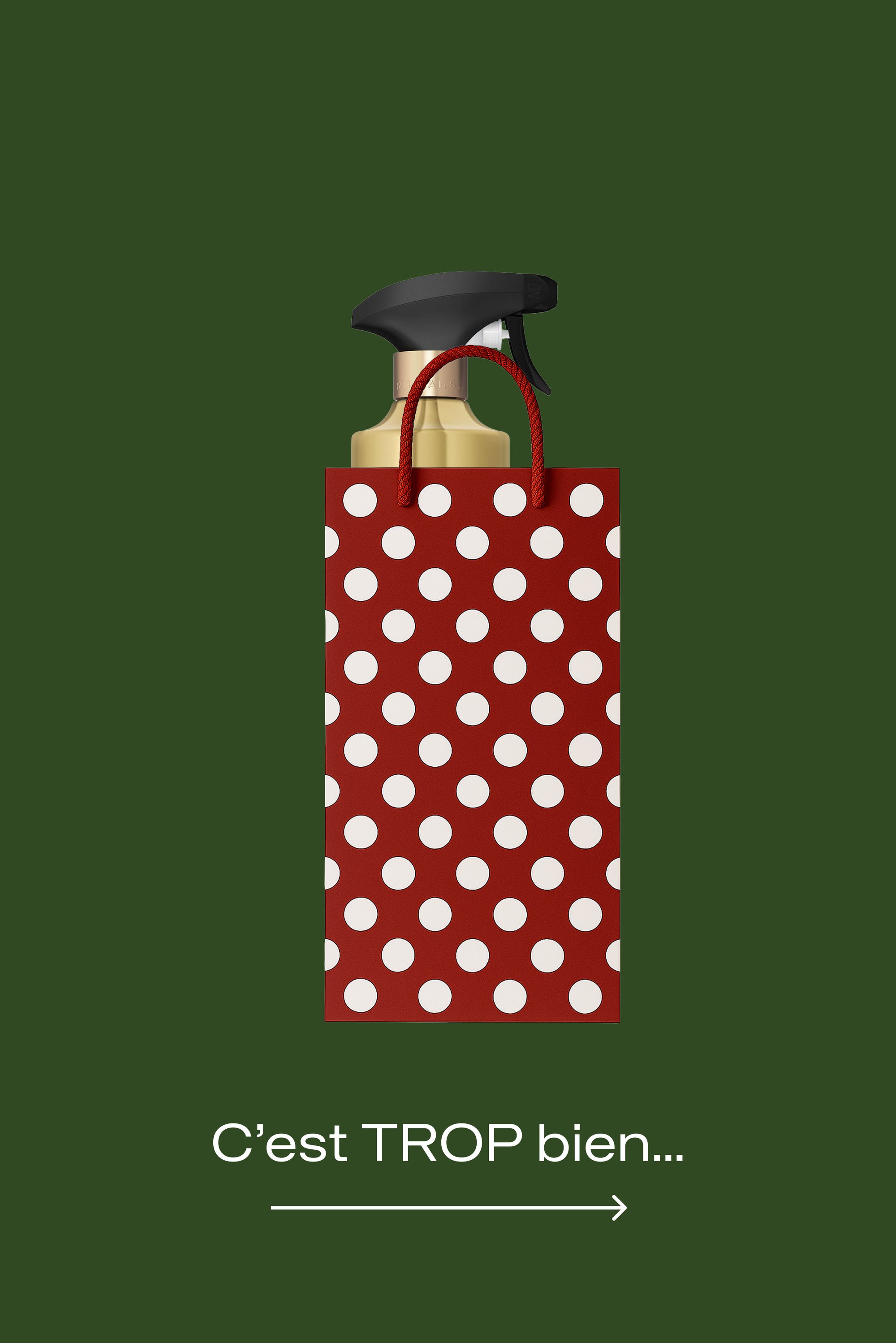 Flacon pulvérisateur doré avec gâchette noire à l'intérieur d'un sac cadeau rouge à pois blancs sur un fond vert foncé avec un texte en français en dessous.