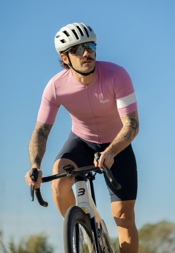 Un cycliste masculin portant un maillot rose et un casque blanc roule à vélo de route sous un ciel bleu clair.