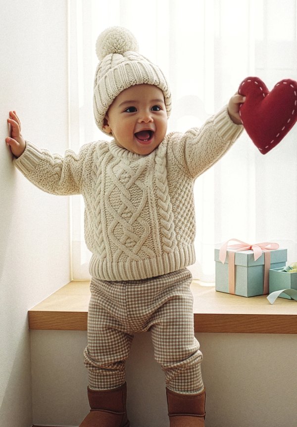 Kleinkind trägt eine Strickmütze und einen Pullover, hält ein rotes Plüschherz und steht an einem Fensterbrett mit pastellfarbenen Geschenkschachteln.