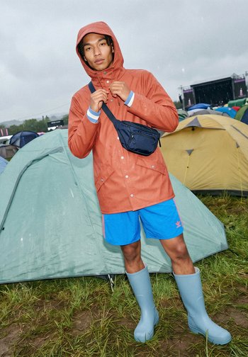 Person trägt orangefarbenen Regenmantel, blaue Shorts und hellblaue Gummistiefel, steht vor Zelten auf nassem Gras bei einer Open-Air-Veranstaltung.