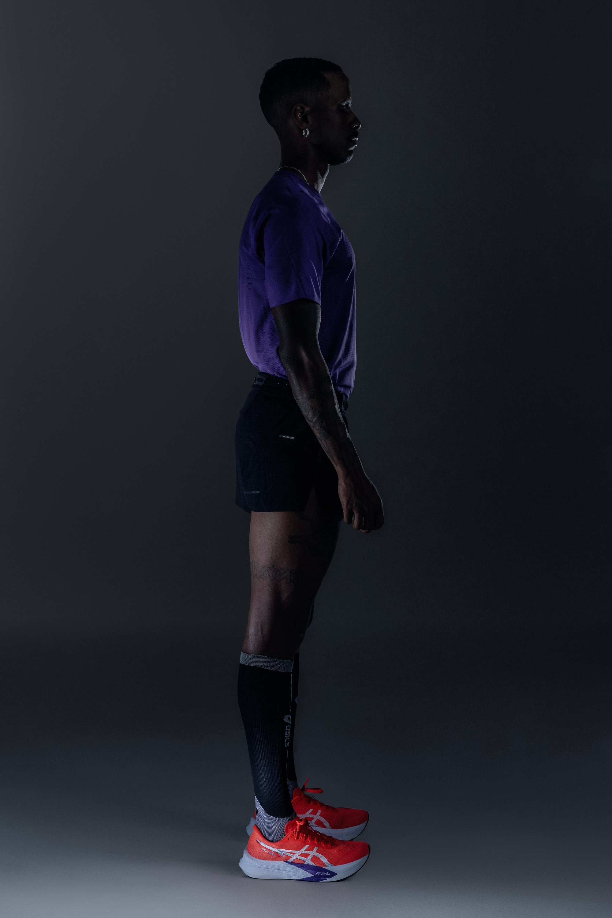 Rote Sportschuhe mit weißen Akzenten, schwarze Shorts und ein lila T-Shirt mit kurzen Ärmeln. Schwarze Kniestrümpfe mit grauen Streifen. Seitenansicht.