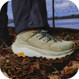 Stivale da trekking su terreno texture con funghi e foglie, sullo sfondo sfocato di una foresta e acqua.