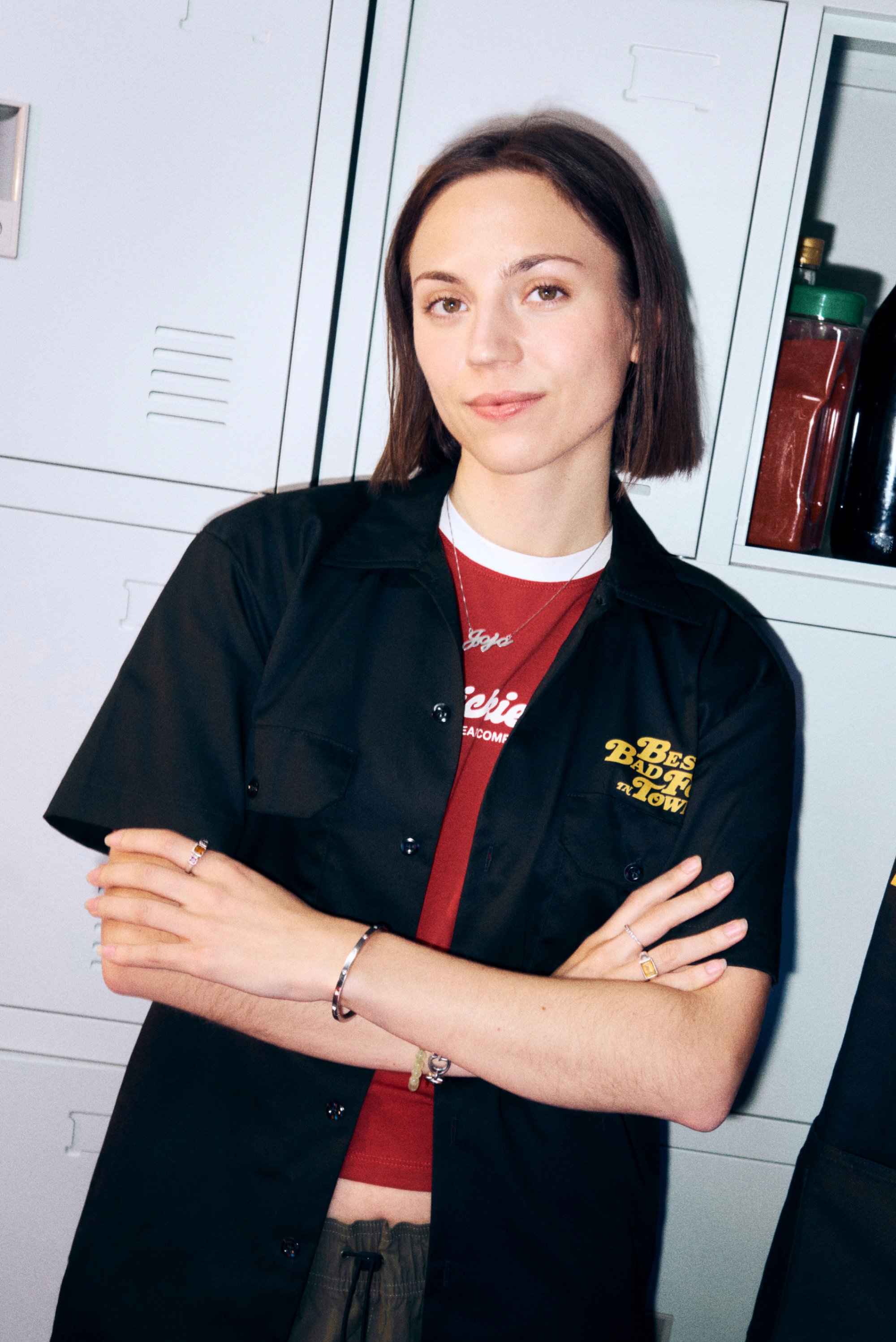 Eine junge Frau mit schulterlangem braunem Haar, die ein schwarzes Shirt und ein rotes Oberteil trägt, posiert mit verschränkten Armen vor Schließfächern.