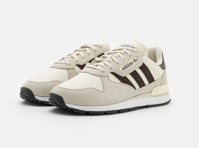 Paire de baskets Adidas beige avec des bandes noires, design à lacets et semelles en caoutchouc, présentées en angle sur fond blanc.