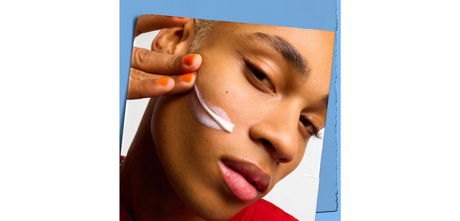 Eine Person, die weiße Creme auf ihre Wange aufträgt, mit kurzen hellen Haaren und orangefarbener Nagellackierung, vor einem blauen Hintergrund.