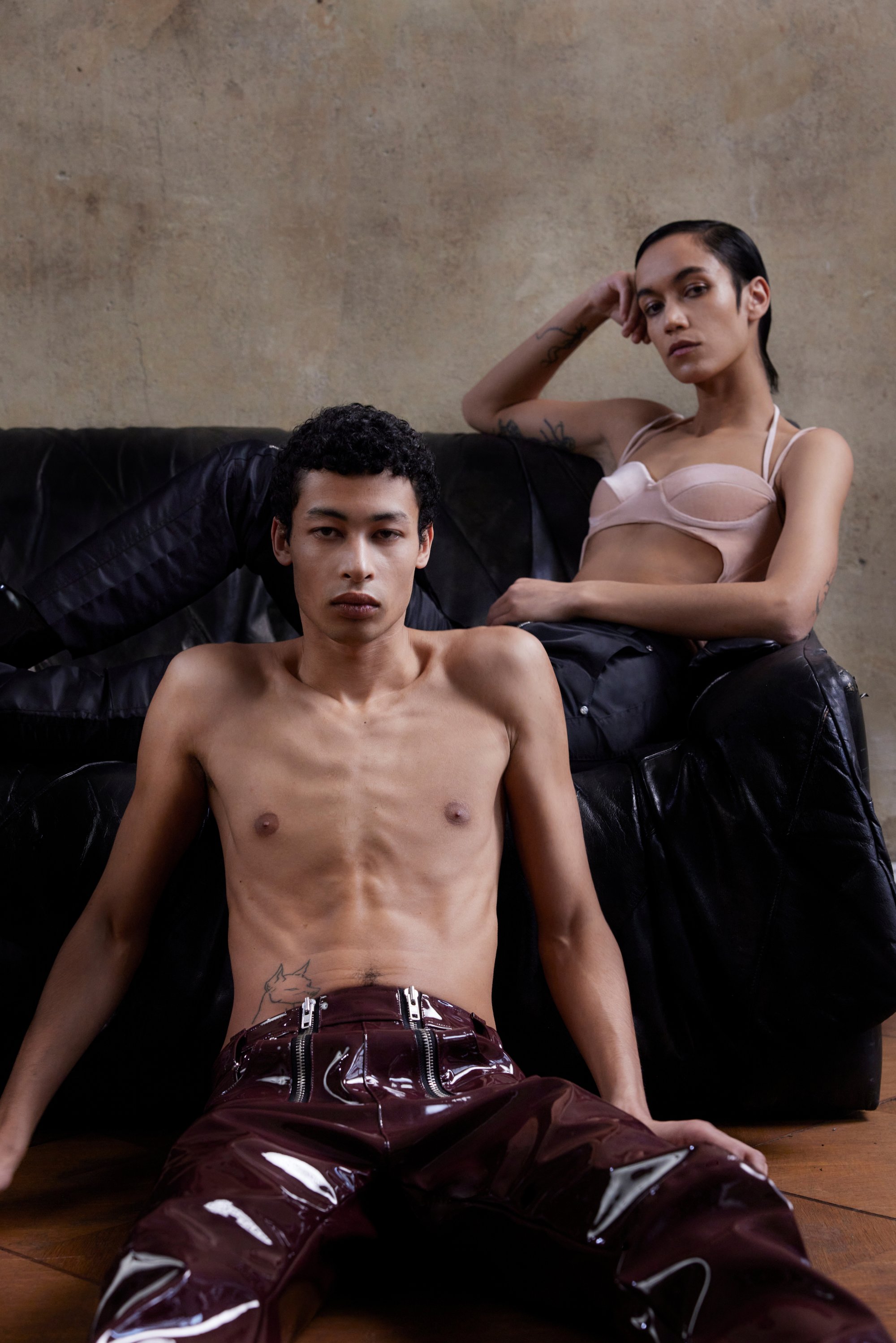 Un giovane senza camicia è seduto sul pavimento in pantaloni in vinile bordeaux, mentre una donna in reggiseno rosa si sdraia su un divano nero.