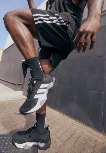 Athlète en shorts et chaussettes noires, équilibrant sur une jambe, mettant en avant des chaussures de sport noir et blanc sur fond urbain.
