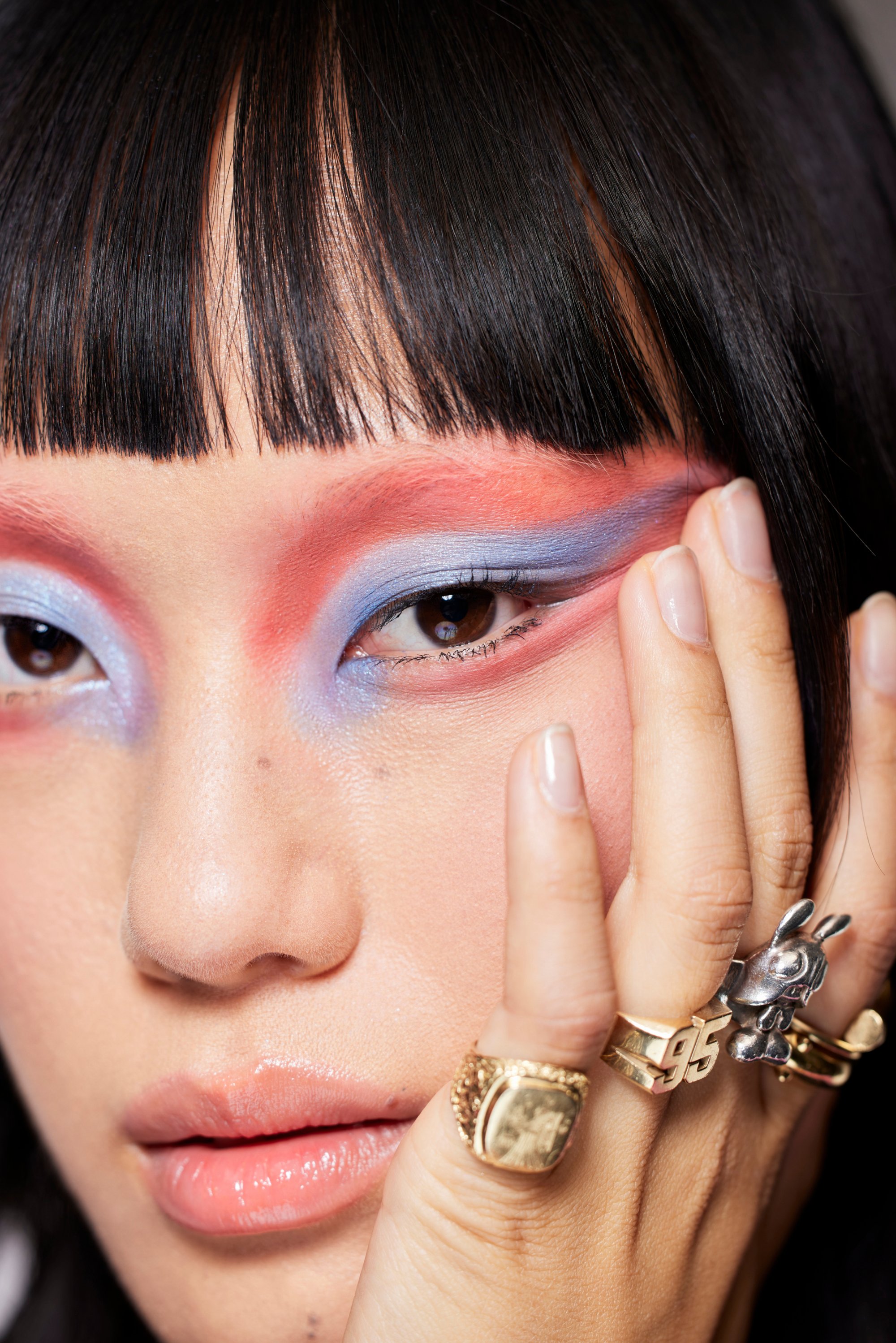 Close-up of a person with bold, colorful eye makeup featuring pink and blue hues, adorned with multiple rings on their hand.
