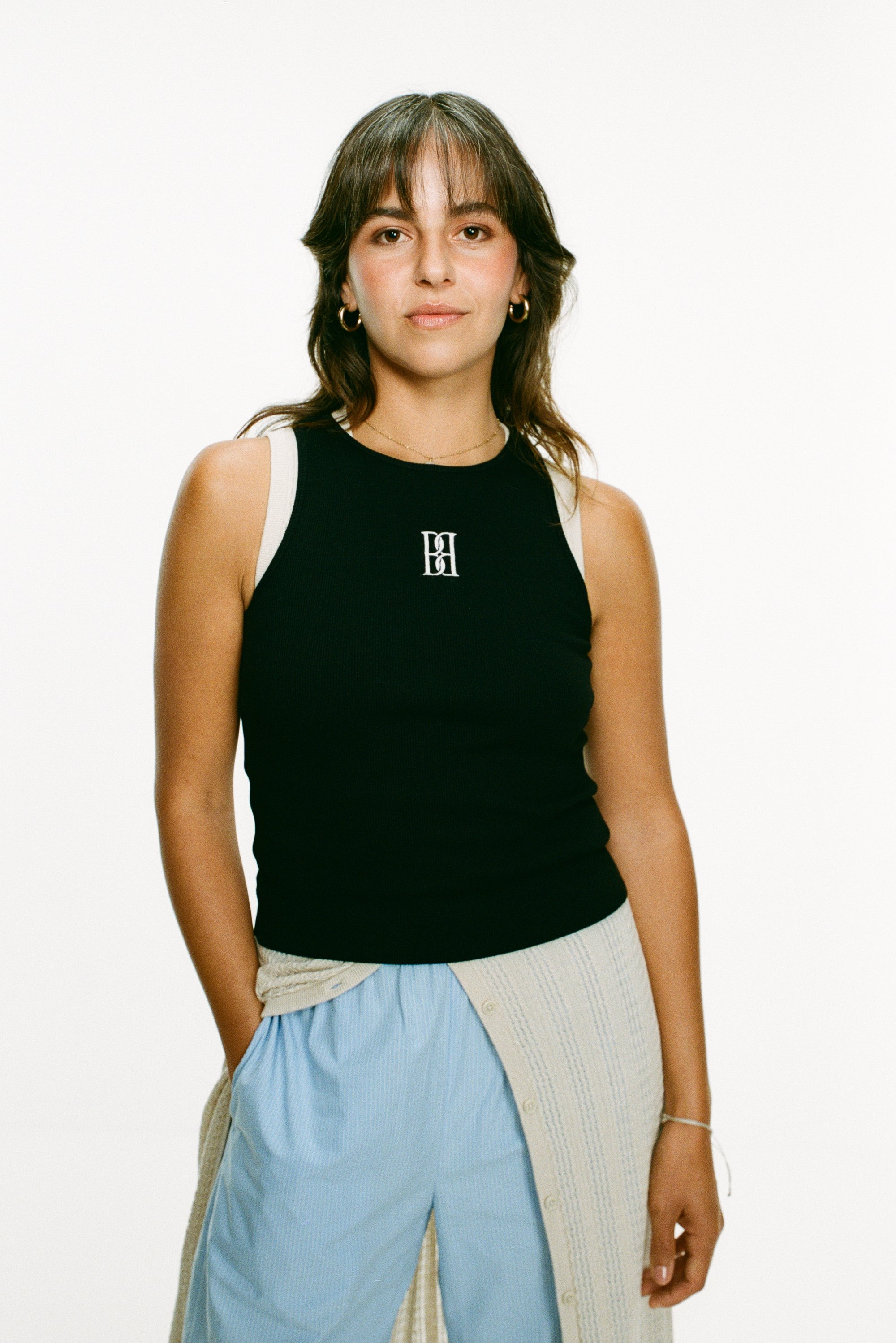 Una donna con capelli castani sta in piedi con sicurezza, indossando una maglietta nera senza maniche con un logo, abbinata a pantaloni blu chiari e un cardigan crema.
