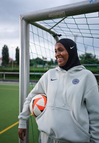 Naeratav naine sportlikus hijabis ja kapuutsiga toetub jalgpallivärava posti vastu, hoides valget ja oranži jalgpalliga rohelisel väljakul.