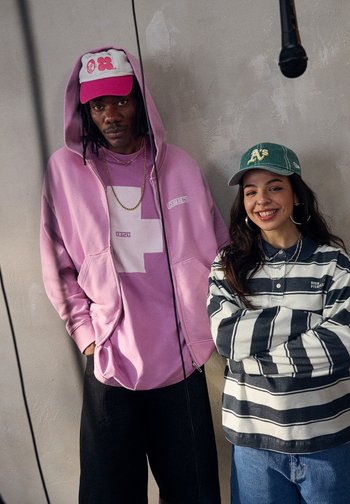 Deux jeunes adultes posent ensemble devant un mur texturé. L'homme porte un hoodie rose et une casquette, tandis que la femme est vêtue d'une chemise rayée et d'une casquette verte.