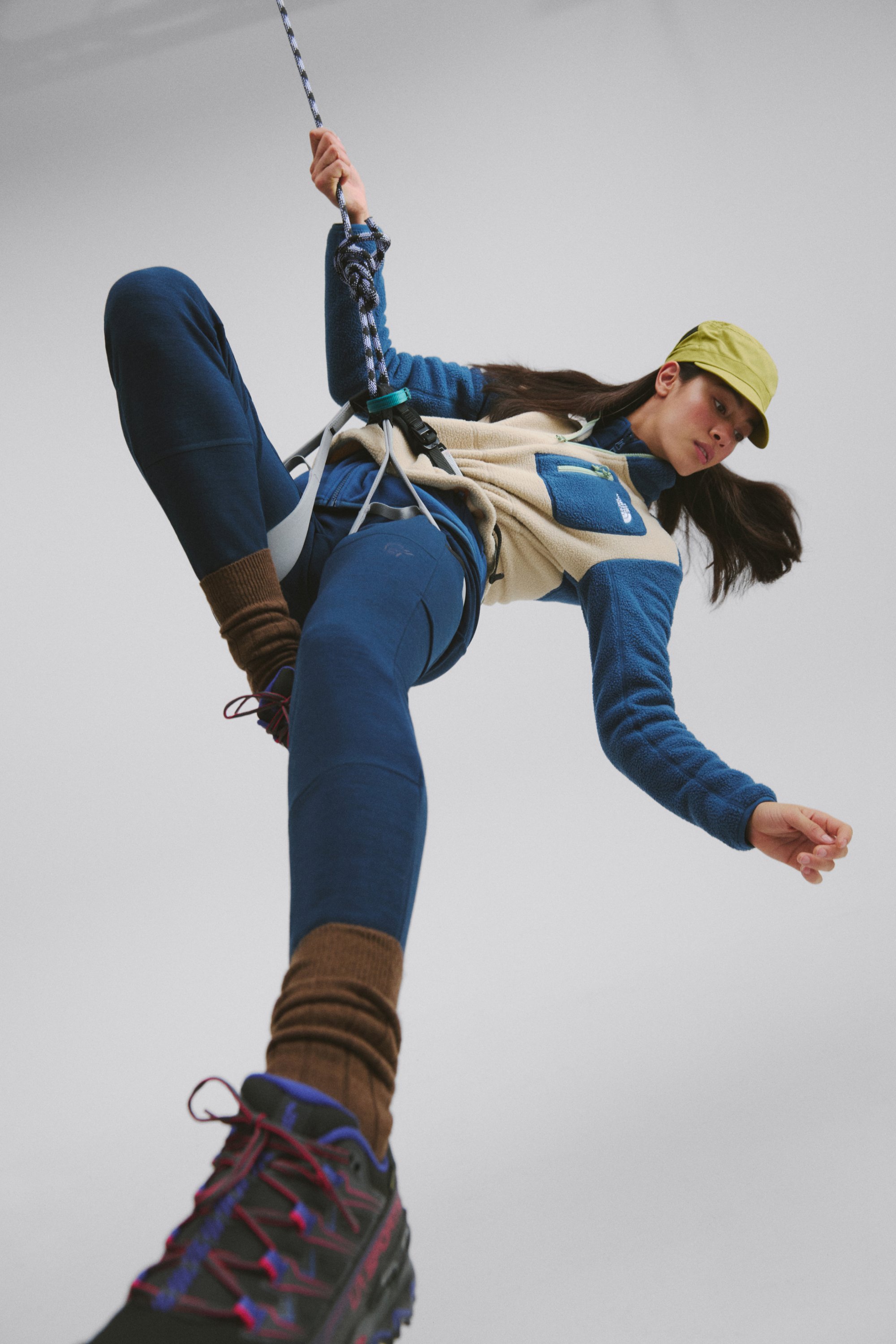 Equipo de escalada en una persona, que lleva un chaleco de forro polar beige con mangas azules, leggins navy, calcetines marrones y zapatos de senderismo negros, agarrando una cuerda.