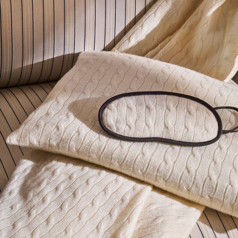 Kabelstrick-Creme-Decke, locker über gestreiftem Stoff drapiert, mit einer schwarzen Schlafmaske darauf liegend. Strukturiertes Muster mit weichem Erscheinungsbild.