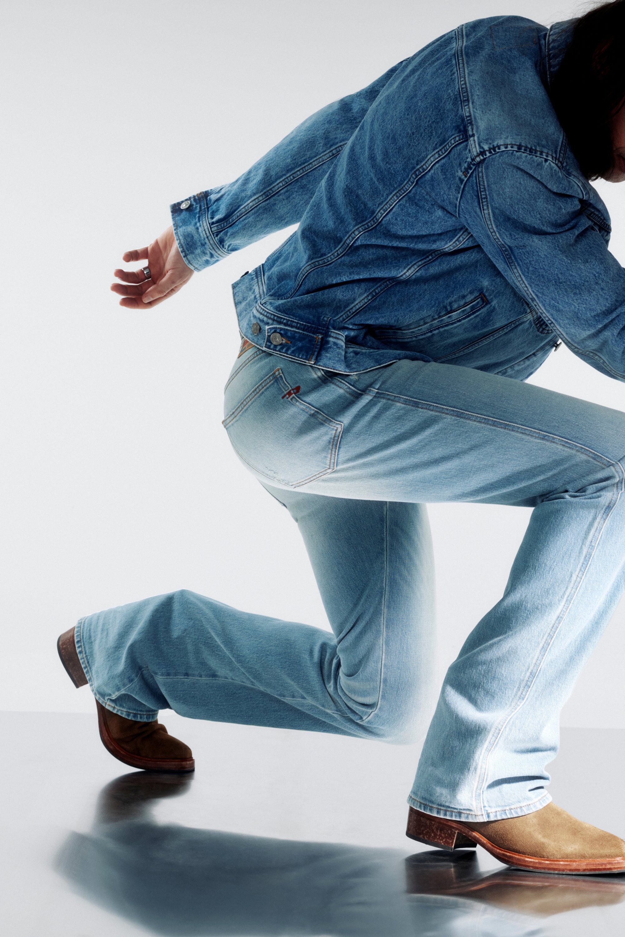 Una persona lleva una chaqueta de mezclilla y unos vaqueros azules acampanados, inclinándose hacia adelante en una pose dinámica sobre una superficie reflectante.