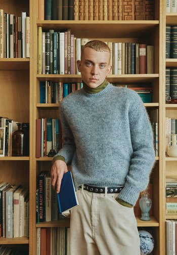 Lichtgrijze, zachte trui met een groene col, gecombineerd met crèmekleurige corduroy broek en een zwarte riem, met een blauw boek in de hand. Boekenplanken op de achtergrond.