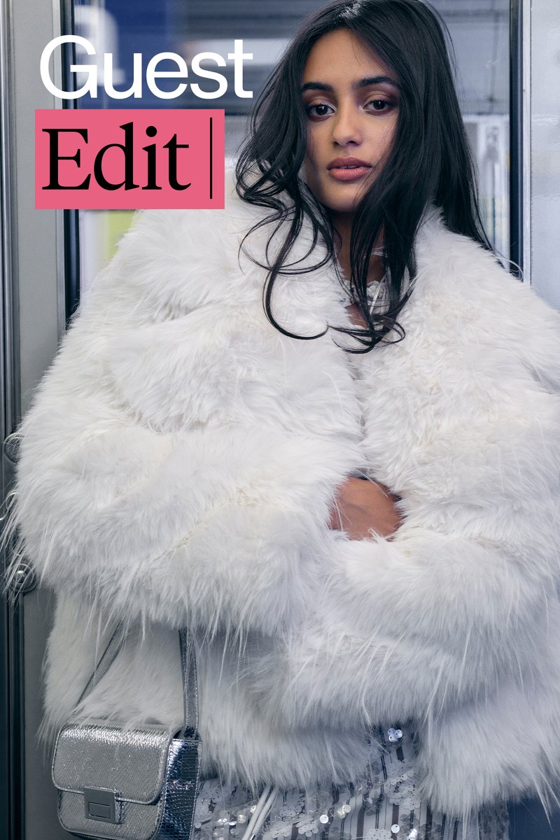 A woman with long dark hair, wearing a fluffy white coat, gazes thoughtfully at the camera in a modern indoor setting.