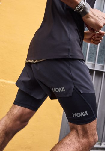 Άνδρας που φοράει μαύρο σορτς για τρέξιμο Hoka και μαύρο στρωματοποιημένο κολάν, σε μέσο βήμα μπροστά σε κίτρινο και γκρι φόντο.