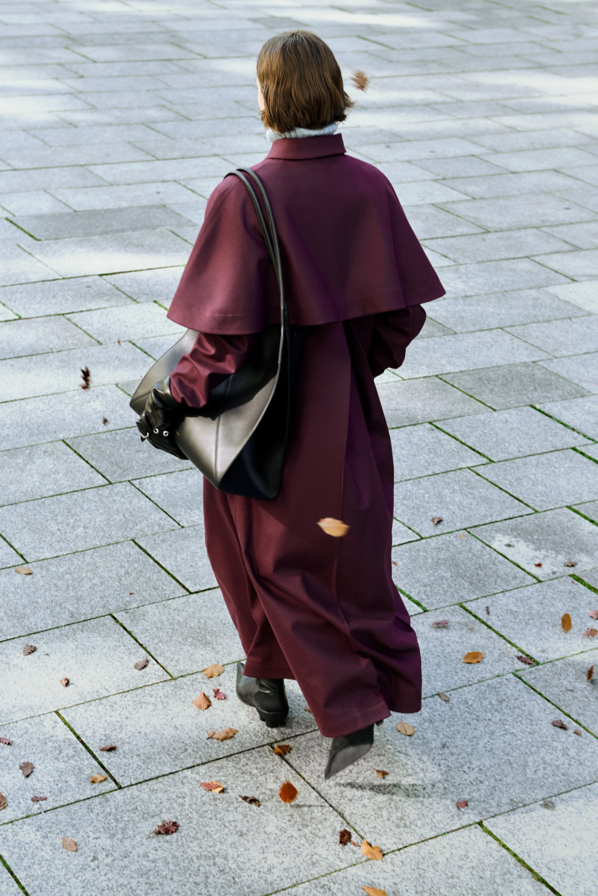 Langer, fließender kastanienbrauner Mantel mit Umlegekragen, kombiniert mit einer schwarzen Handtasche und Stiefeln. Betonpflaster mit verstreuten Blättern.