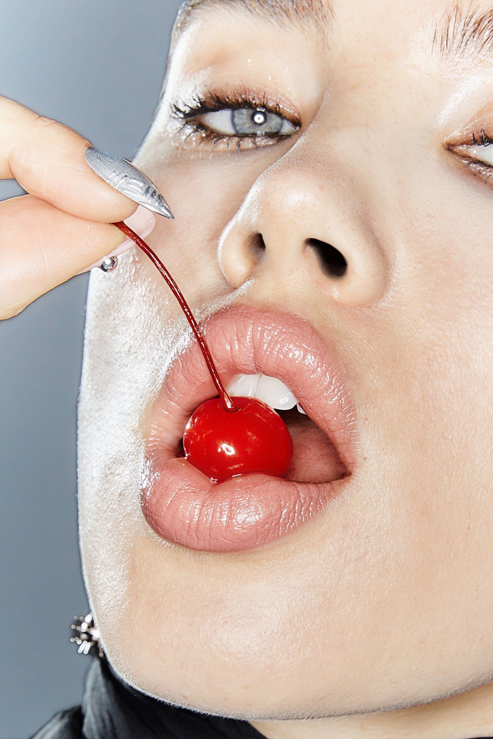 Close-up van een persoon die een kers met een steeltje bij hun lippen vasthoudt, waarbij glanzende huid en zachte make-up worden getoond tegen een neutrale achtergrond.