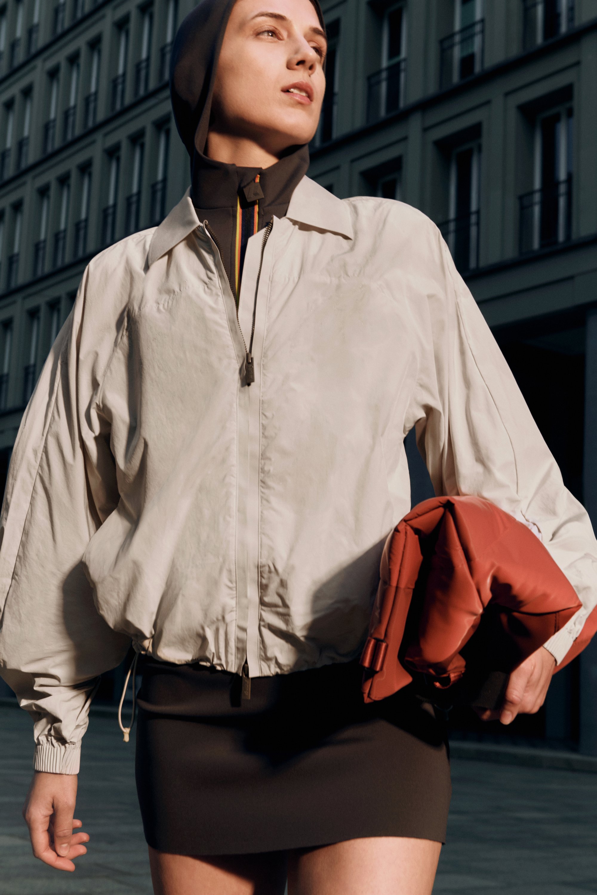 Helle beigefarbene Bomberjacke mit einem lässigen Schnitt, detaillierten Reißverschlüssen und einem braunen Rock. Hält eine rote gepolsterte Tasche mit abgerundeten Kanten.