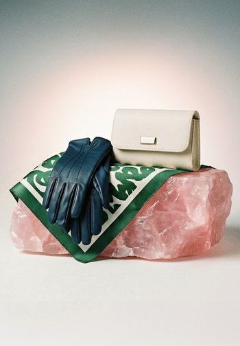 Melnas ādas cimdi un krēmkrāsas soma, kas atrodas uz zaļas rakstainas šalles virs liela rozā kristāla akmens.