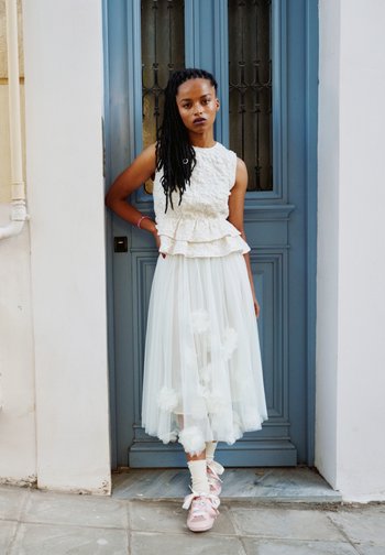 Woman with long dark hair in a white textured sleeveless top and tulle skirt with floral details, standing against a blue door with one hand on hip.