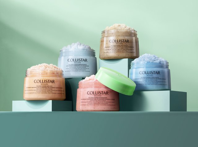 Five jars of COLLISTAR body scrubs in various colors (pink, blue, green, beige) are arranged on light teal blocks against a pale green backdrop.