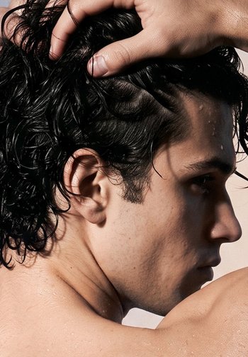 Young man with wet dark curly hair holds head with hand, face in profile, water droplets visible on skin and hair.
