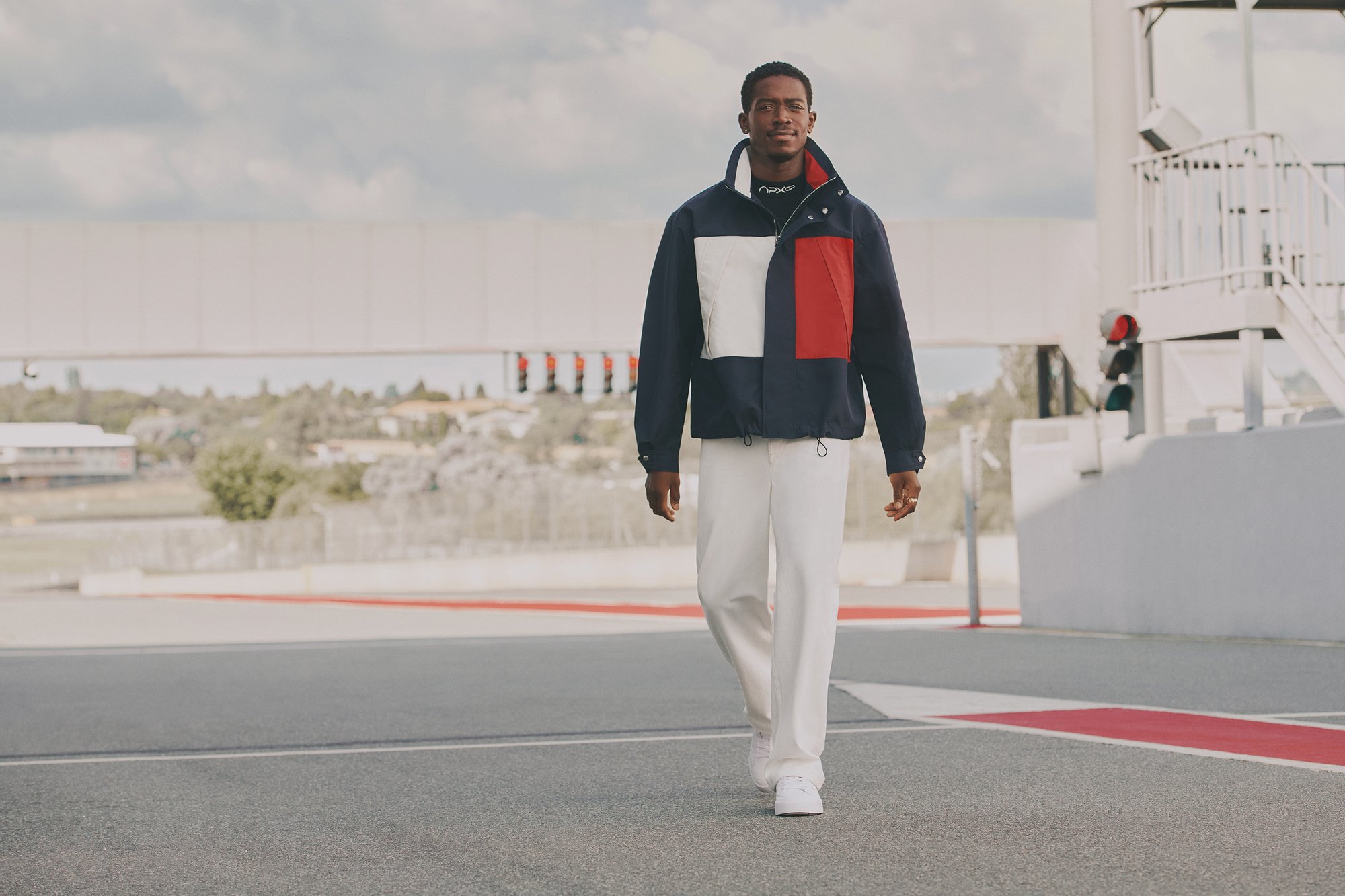 Un hombre camina con confianza en una pista de carreras, vistiendo una chaqueta a bloques de color, pantalones blancos y zapatillas, con un cielo nublado de fondo.
